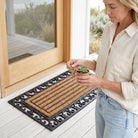 Woman holding keys and a small plant on a decorative doormat with a wooden door in the background.