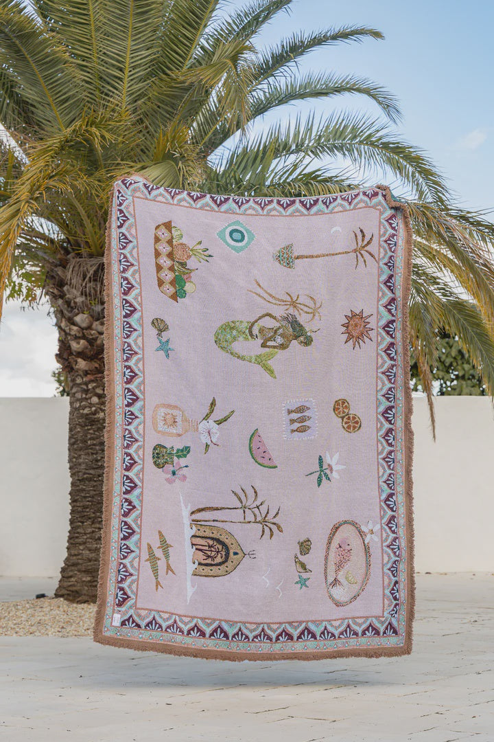 Decorative throw with mermaid and shell patterns hanging against a palm tree and white wall background.