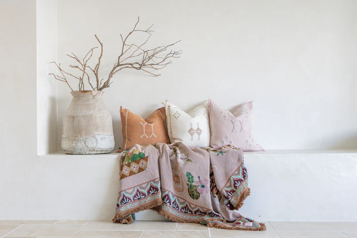 Decorative pillows and throw blanket on a bench with a vase and branches in the background.