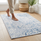 A light blue and off-white patterned area rug on a light wood floor, with a person's bare feet walking on it. A woven basket and a potted plant are visible in the background near a window.