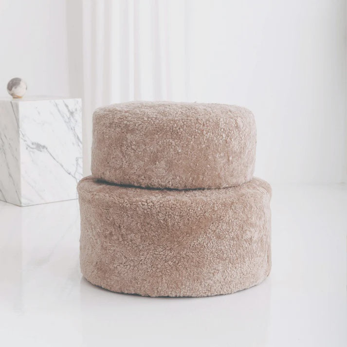 Two fluffy beige ottomans on a white surface with a marble box in the background.