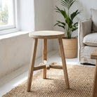A light natural wood barrel stool with a round seat and four angled legs, shown in a bright, modern room on a woven rug next to a sofa and potted plant.