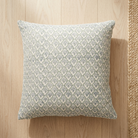 A square throw pillow with a repeating blue-gray and off-white floral pattern, resting on a light wood plank floor next to a woven jute rug.