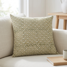A square throw pillow with an olive green and beige geometric leaf pattern, resting on a light beige fabric sofa in a bright, modern living room setting.