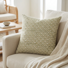 A square decorative throw pillow with an olive green and cream geometric floral pattern, resting on a light beige linen armchair in a bright, modern living room setting.