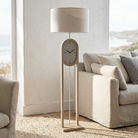 A modern floor lamp with a cylindrical beige fabric shade, a central oval clock face, and a slim rectangular wooden base, styled in a bright living room next to a beige sofa.