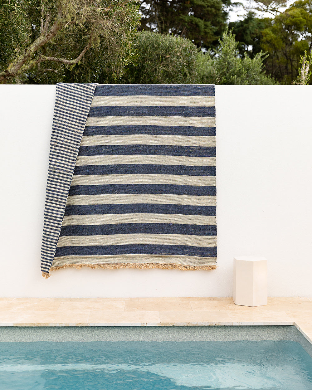 A striped woven textile, likely a beach towel or blanket, draped over a white wall above a pool. The fabric features alternating navy blue and beige horizontal stripes with a fringed edge, set against a background of green trees and a beige stone deck.