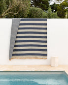 A striped woven textile, likely a beach towel or blanket, draped over a white wall above a pool. The fabric features alternating navy blue and beige horizontal stripes with a fringed edge, set against a background of green trees and a beige stone deck.