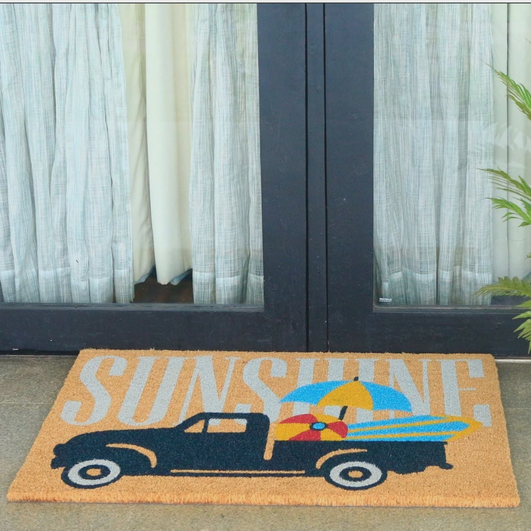 A rectangular coir doormat with a vintage pickup truck carrying surfboards and a beach ball, with the word 'SUNSHINE' in large letters across the top, placed in front of glass patio doors with curtains.