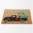 A rectangular coir doormat featuring a black pickup truck carrying a blue, yellow, and red beach umbrella and surfboards, with the word 'SUNSHINE' in large light gray letters across a natural tan background.