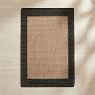 A rectangular area rug with a natural tan woven center and a contrasting black textured border, laid flat on a light wood floor with soft leaf shadows.
