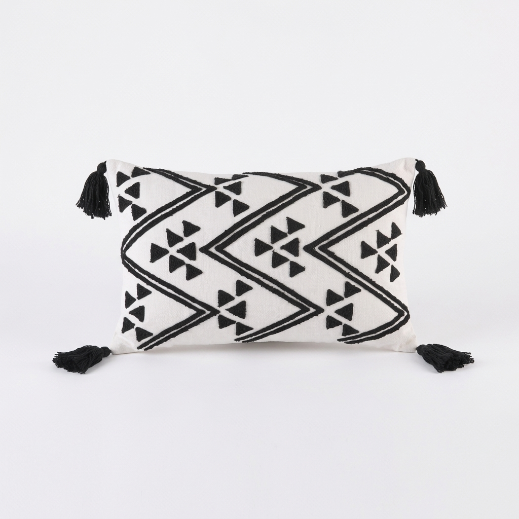 A square throw pillow with a black and white geometric pattern and black tassels on all four corners, photographed against a seamless white background.