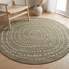 A round area rug with a textured olive green base and a white dotted spiral pattern, styled on a light wood floor in a bright room next to a wooden chair and a vase.