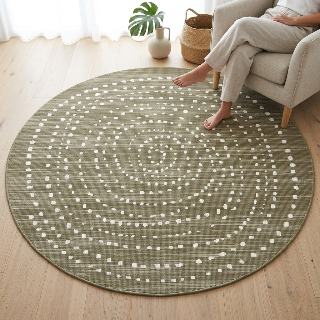 A round area rug with a textured olive green base and a white dotted spiral pattern, shown on a light wood floor in a living room setting with a person sitting on a beige armchair.