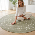 A woman sits cross-legged on a round sage green area rug with a white dotted geometric pattern, placed on a light wood floor in a bright, modern room.