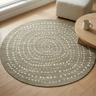 A round area rug with a textured taupe-gray base and a white dotted spiral pattern, styled on a light wood floor next to a wooden coffee table and a light beige ottoman.