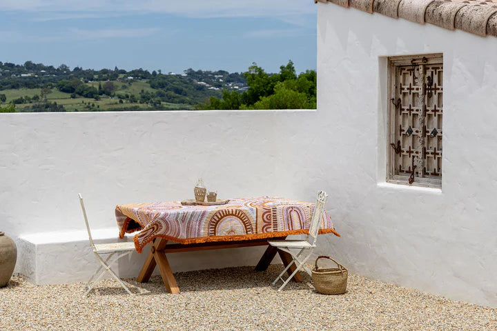 Outdoor setting with a table draped with a colorful table throw, chairs, and a woven basket against a white wall with a window.