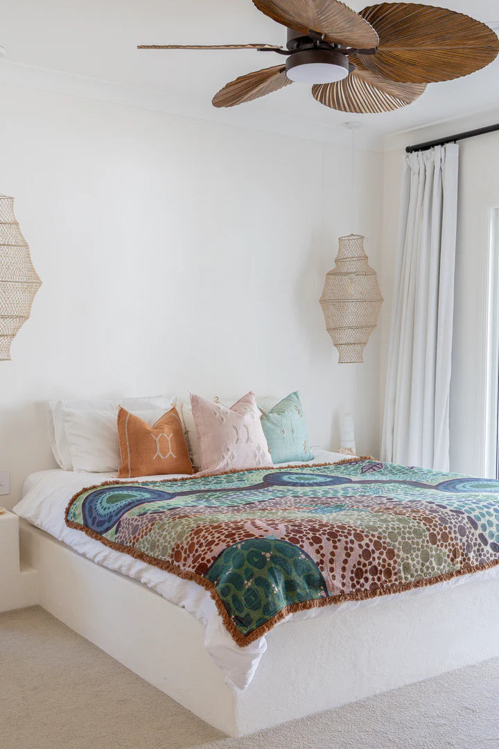 Bedroom with a bed featuring a colorful blanket and pillows, wooden ceiling fan, and decorative lamps.