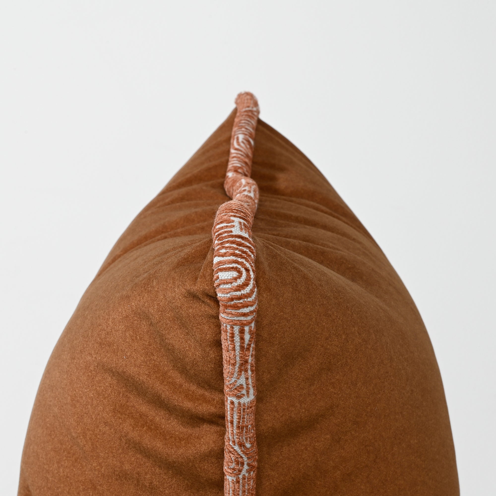 A close-up of a brown textured throw pillow with a decorative vertical tufted trim featuring a white and terracotta pattern, set against a plain light gray background.