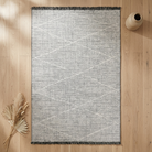 A rectangular area rug with a textured gray and off-white geometric diamond pattern, styled on a light wood floor with dried pampas grass and a ceramic vase.