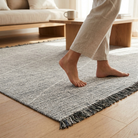 A close-up of a person's bare feet stepping onto a textured gray and white woven area rug with black fringe, placed on a light wood floor in a bright, modern living room.
