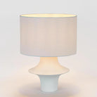 A modern table lamp with a textured light beige fabric drum shade and a sculptural white ceramic base, shown illuminated against a clean white background.