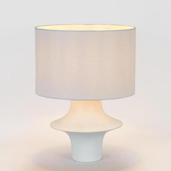 A modern table lamp with a textured light beige fabric drum shade and a sculptural white ceramic base, shown illuminated against a clean white background.