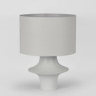 A modern table lamp with a textured light gray fabric drum shade and a sculptural, tiered white ceramic base, shown against a neutral light gray background.