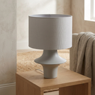 A modern table lamp with a textured light gray fabric drum shade and a matching light gray sculptural base, shown sitting on a wooden side table in a softly lit room.