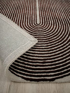 A close-up of a patterned area rug with concentric wavy stripes in dark brown and cream, with a folded corner showing the textured cream backing and edge.