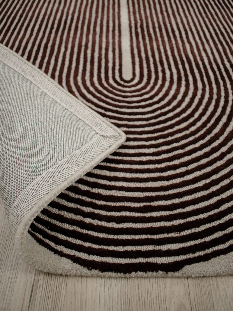 A close-up of a patterned area rug with concentric wavy stripes in dark brown and cream, with a folded corner showing the textured cream backing and edge.