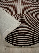 A close-up of a patterned area rug with concentric wavy stripes in dark brown and cream, with a folded corner showing the textured cream border, placed on a light wood floor.