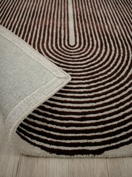 A close-up of a patterned area rug with concentric wavy stripes in dark brown and cream, with a folded corner showing the textured cream border, placed on a light wood floor.