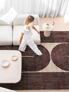 A child in a white outfit walks barefoot across a large patterned area rug in a bright, modern living room with a white sofa, low tables, and sheer curtains.