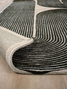 A close-up of a modern area rug with a dark charcoal and cream wavy geometric pattern, shown with one corner rolled up to reveal the backing and edge, placed on a light wood floor.