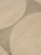 A close-up of a textured area rug featuring a modern abstract pattern of concentric wavy lines in warm beige and taupe-gray.