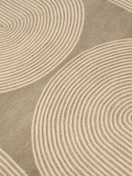 A close-up of a textured area rug featuring a modern abstract pattern of concentric wavy lines in warm beige and taupe-gray.