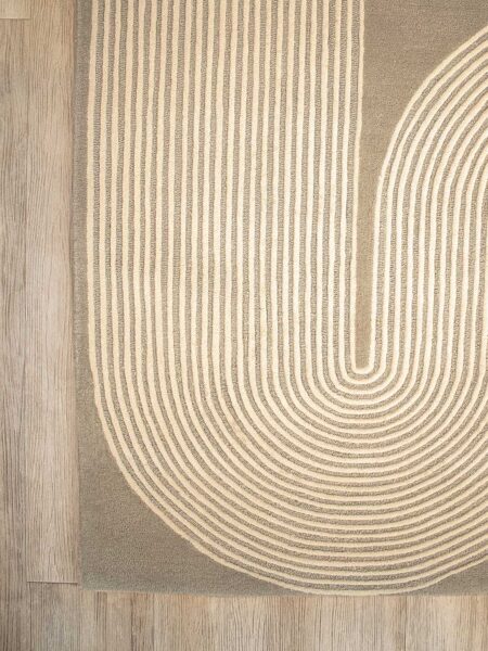 A close-up of a rug with a geometric pattern of concentric arches in cream on a taupe background, shown on a light wood floor.