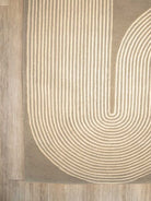 A close-up of a rug with a geometric pattern of concentric arches in cream on a taupe background, shown on a light wood floor.