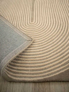 A close-up of a textured area rug with concentric curved stripes in beige and taupe, showing the underside with a woven gray backing and a folded corner revealing the wood floor underneath.