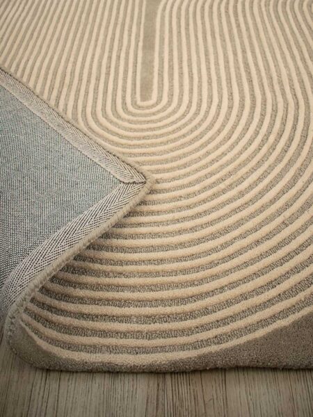 A close-up of a textured area rug with concentric curved stripes in beige and taupe, showing the underside with a woven gray backing and a folded corner revealing the wood floor underneath.