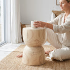 A woman in light beige loungewear sits cross-legged on a woven jute rug next to a small round wooden side table, holding a white mug in a bright, modern room.
