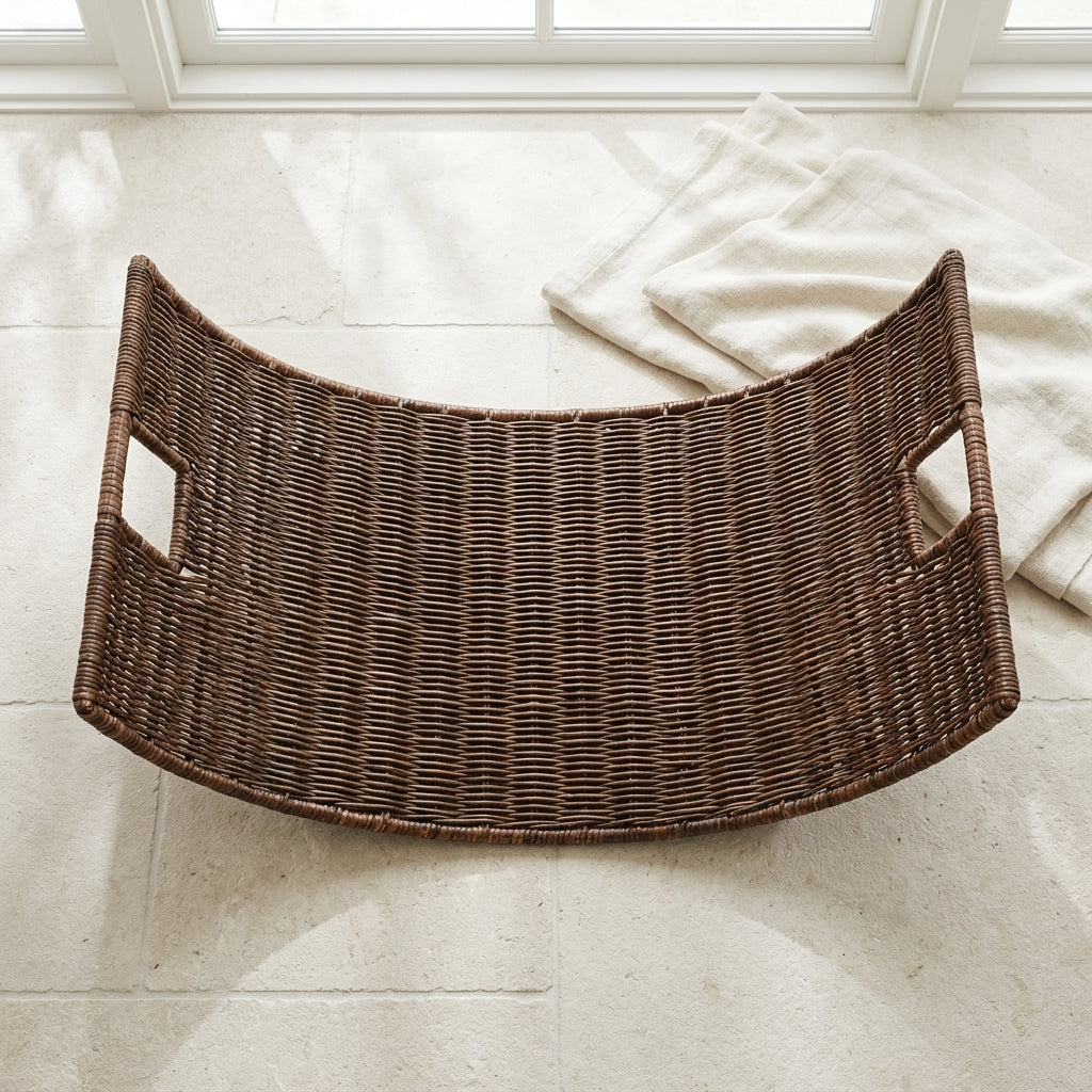 A curved dark brown woven rattan tray with two side handles, placed on a light beige tiled floor near a window with a folded cream towel nearby.