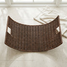 A curved dark brown woven rattan tray with two side handles, placed on a light beige tiled floor near a window with a folded cream towel nearby.