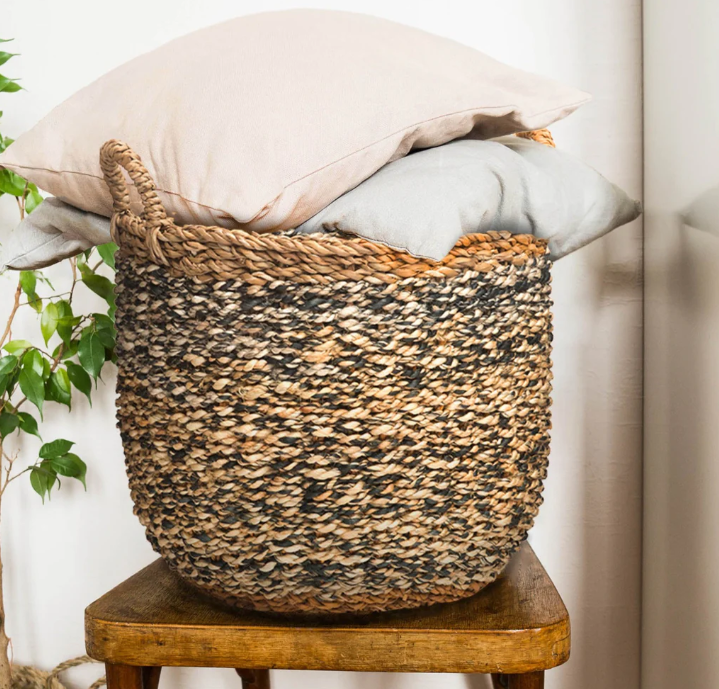 Woven basket with pillows on a wooden stool against a neutral wall.