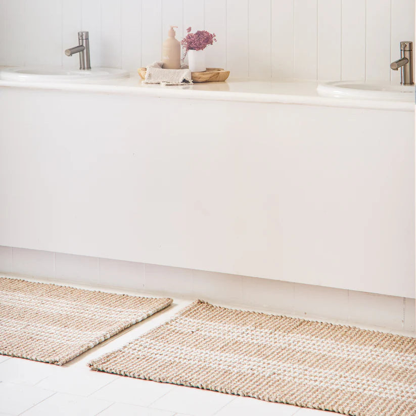Two woven bath mats on a bathroom floor with a white bathtub and sink in the background.