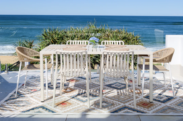 Outdoor dining set with white table and chairs on a patio by the ocean.