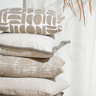 A stack of four decorative throw pillows in neutral beige and white tones, including a patterned pillow on top, set against a soft white curtain background with dried palm leaves in the corner.