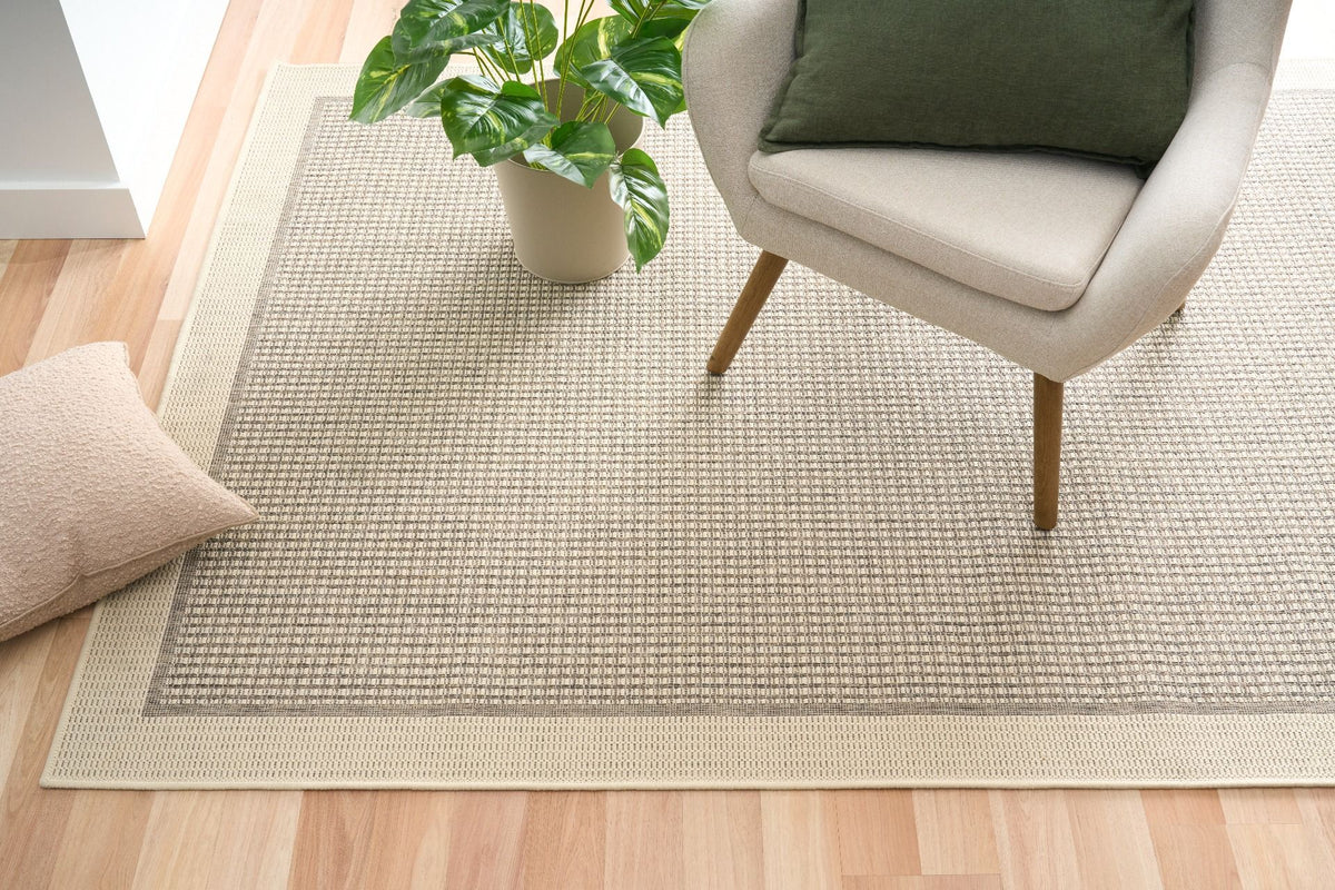 Beige woven rug on a wooden floor with a chair and plant in the background