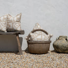 Two beige and cream patterned throw pillows with fringed edges, styled on a rustic wooden bench and in a woven basket against a white stucco wall on a gravel surface.